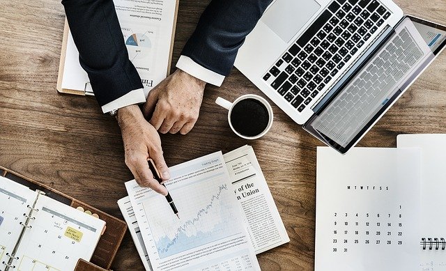 a male MROC manager pointing at chart with a laptop, a calendar, and a planner all on top of the desk